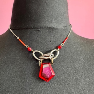 May include: A silver-toned necklace with a large, faceted, red gemstone pendant. The necklace features a delicate chain, red bead accents, and a geometric design. The pendant is a vibrant red color with a textured interior.