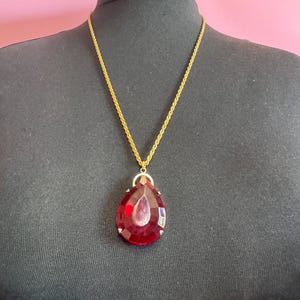 May include: A vintage gold-coloured necklace featuring a large, teardrop-shaped pendant. The pendant is a deep red colour with a faceted design, suspended from a delicate gold chain. The necklace is displayed on a black mannequin.