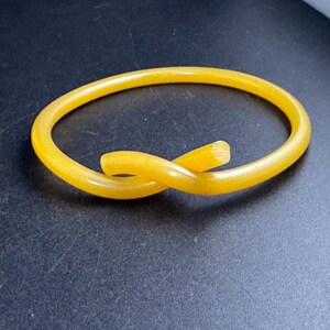 May include: A yellow bangle bracelet with a knotted design. The bracelet is made of a solid, translucent material and has a smooth, polished finish. The bracelet is circular and appears to be approximately 6.35 cm in diameter.