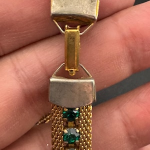 May include: Close-up of a vintage gold-tone bracelet with emerald green gemstones. The bracelet features a mesh chain design and rectangular silver-tone clasps. The gemstones are round and set in a row.