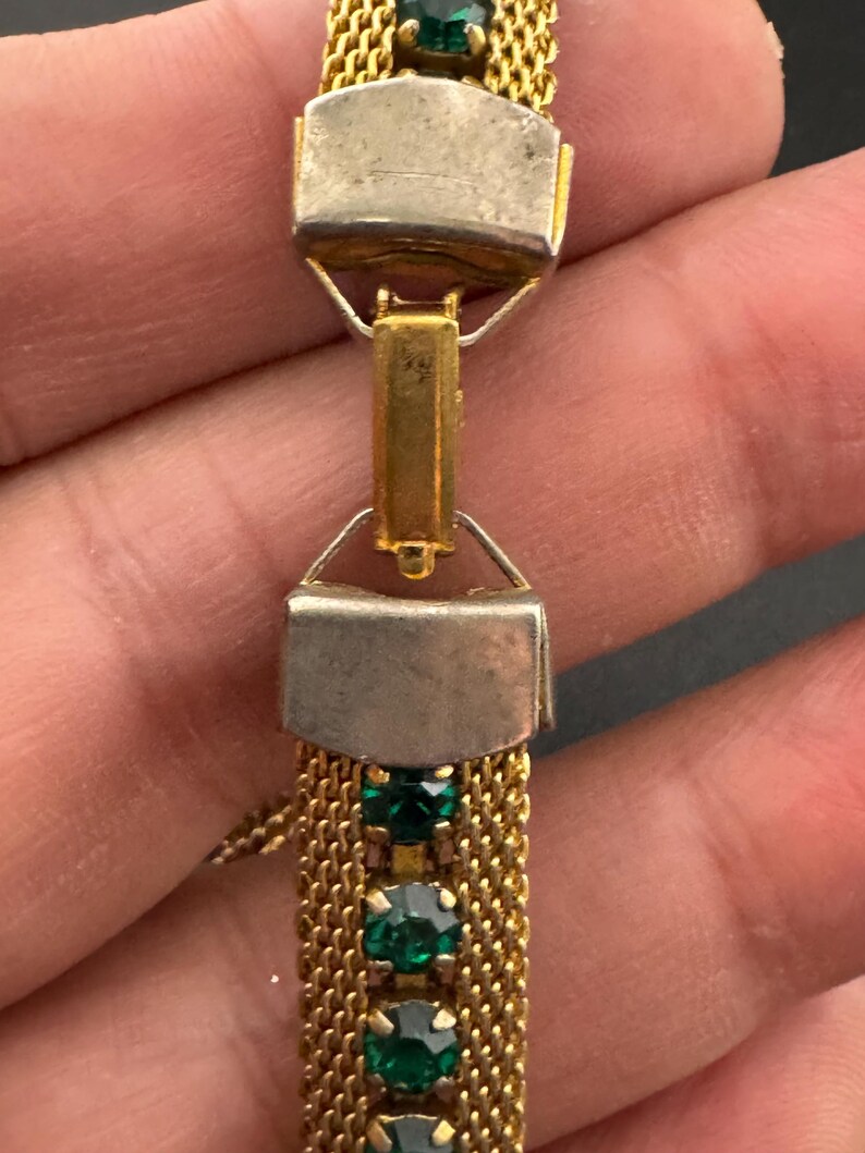 May include: Close-up of a vintage gold-tone bracelet with emerald green gemstones. The bracelet features a mesh chain design and rectangular silver-tone clasps. The gemstones are round and set in a row.