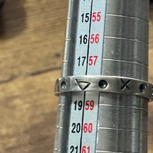 Może przedstawiać: Srebrny pierścionek z tr&oacute;jkątem, k&oacute;łkiem i literą X. Pierścionek jest mierzony na metalowym mierniku pierścionk&oacute;w z oznaczeniami od 14 do 22.