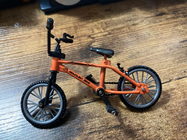 Op de afbeelding: Miniatuur oranje mountainbike met zwarte banden, zadel en stuur. De fiets heeft de woorden "Mountain Bike" op het frame gedrukt. De wielen hebben zilveren spaken. Een kleine waterfles is aan het frame bevestigd.