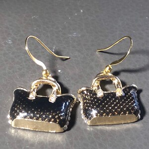 Puede incluir: Un par de pendientes colgantes de tono dorado con un dise&ntilde;o de bolso negro y detalles de diamantes de imitaci&oacute;n transparentes.