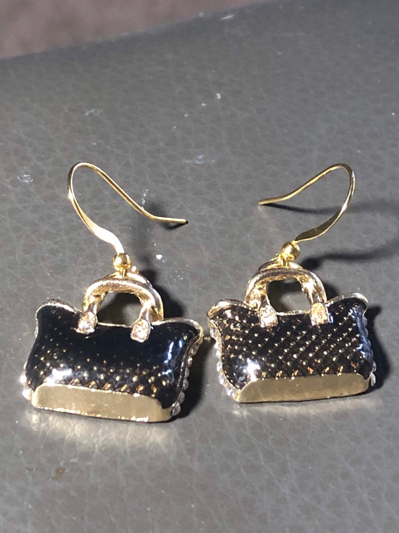 Puede incluir: Un par de pendientes colgantes de tono dorado con un dise&ntilde;o de bolso negro y detalles de diamantes de imitaci&oacute;n transparentes.