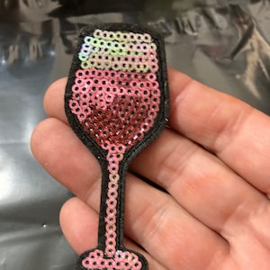 Peut inclure: Un patch en forme de verre à vin en sequins rose et blanc. Le verre est rempli d'un liquide rose et a un dos en feutre noir.