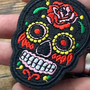 May include: Embroidered black sugar skull patch with colourful details, including a red rose, yellow and white circles, and pink teeth.