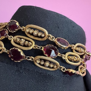 May include: A gold-tone metal chain bracelet featuring alternating oval links with textured edges and small central studs, interspersed with faceted deep red glass stones. The bracelet is constructed with multiple connected segments.