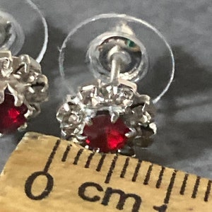 Puede incluir: Un par de pendientes de plata con un centro de cristal rojo y un halo de cristal transparente. Los pendientes tienen un di&aacute;metro de 1 cm.