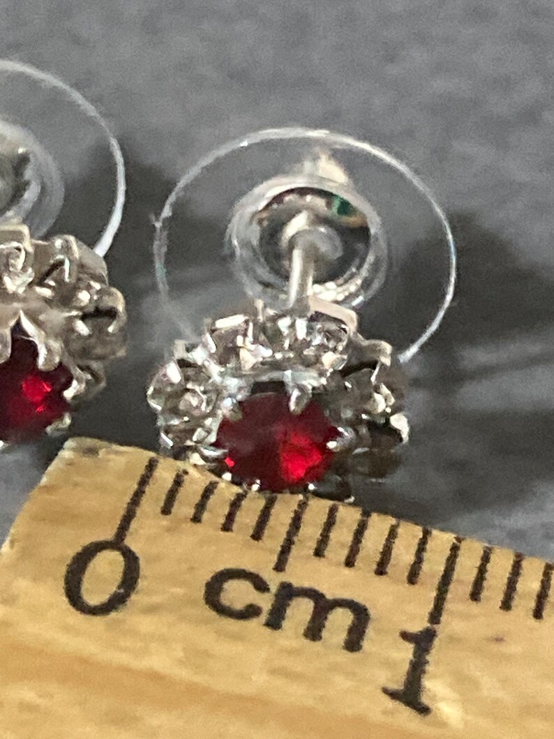 Puede incluir: Un par de pendientes de plata con un centro de cristal rojo y un halo de cristal transparente. Los pendientes tienen un di&aacute;metro de 1 cm.