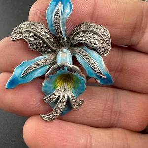 May include: A vintage silver-tone brooch in the shape of an orchid. The brooch features light blue enamel petals with silver-tone detailing and a yellow center. The brooch is a floral accessory.