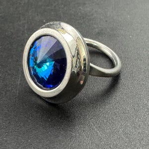 May include: A silver ring featuring a large, round, faceted blue gemstone. The gemstone is set in a bezel setting, and the ring band is a simple, smooth design. The ring is displayed on a dark surface.
