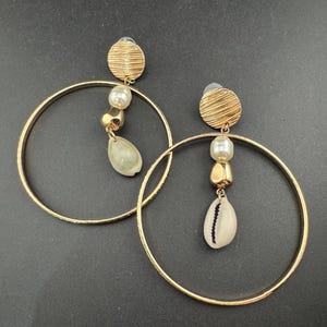 May include: A pair of gold-coloured hoop earrings. Each earring features a textured gold disc, a pearl, a gold bead, and a white seashell pendant. The earrings are displayed against a dark background.