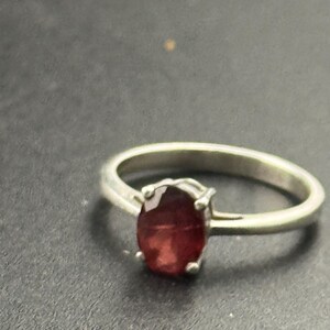 May include: A silver ring featuring a deep red oval gemstone. The ring's band is a simple, polished silver, and the gemstone is held in place by four prongs. The ring is set against a dark background.