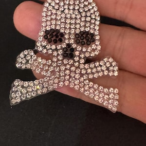 以下が含まれることがあります： キラキラ光るラインストーンがちりばめられた、スカルとクロスボーンのデザイン。スカルの眼窩は黒い石で埋められ、印象的なコントラストを生み出しています。装飾用のパーツです。