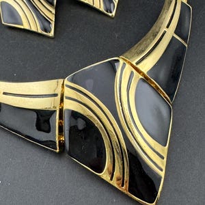 May include: A vintage jewelry set featuring a necklace and matching earrings. The set has a gold-tone metal base with black enamel geometric shapes and gold accent lines. The earrings are square-shaped, and the necklace has a bib design.