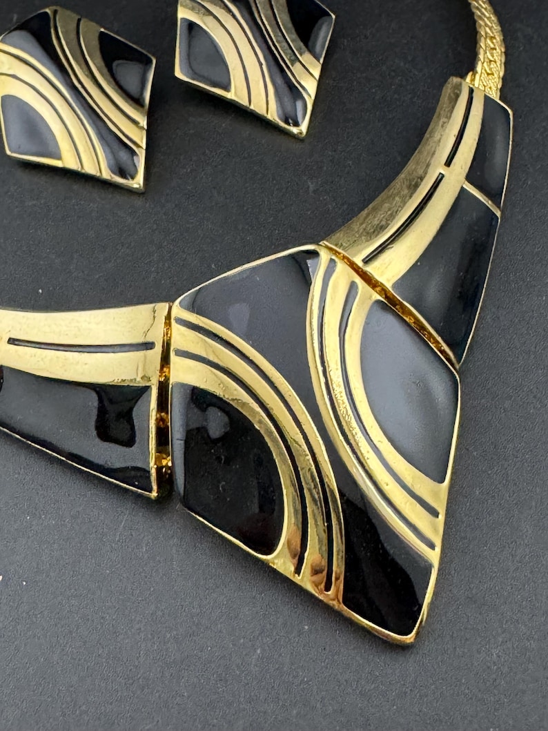May include: A vintage jewelry set featuring a necklace and matching earrings. The set has a gold-tone metal base with black enamel geometric shapes and gold accent lines. The earrings are square-shaped, and the necklace has a bib design.
