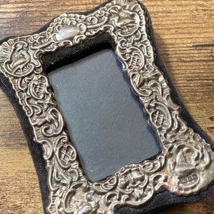Puede incluir: Un marco de fotos de plata con un dise&ntilde;o ornamentado y un respaldo de terciopelo negro. El marco tiene una peque&ntilde;a inscripci&oacute;n en la parte inferior que dice "KFD".