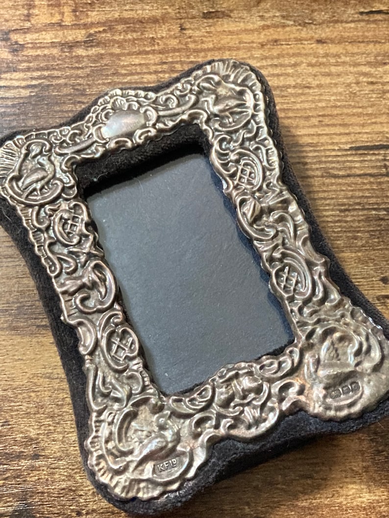 Puede incluir: Un marco de fotos de plata con un dise&ntilde;o ornamentado y un respaldo de terciopelo negro. El marco tiene una peque&ntilde;a inscripci&oacute;n en la parte inferior que dice "KFD".