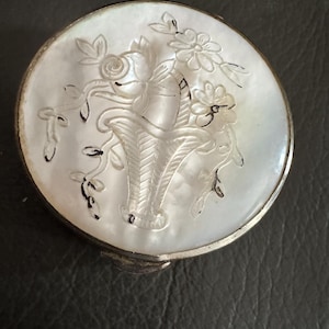 May include: A round, silver-toned metal compact case featuring an iridescent white mother-of-pearl inlay on the lid. The inlay is intricately carved with a floral basket design, with dark pigment highlighting the engraved lines of the flowers.