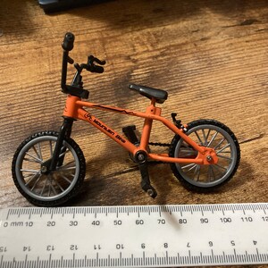 Op de afbeelding: Een oranje en zwarte miniatuur mountainbike met de woorden "Mountain Bike" op het frame. De fiets heeft zilveren wielen en een zwart zadel. Een liniaal staat op de voorgrond.