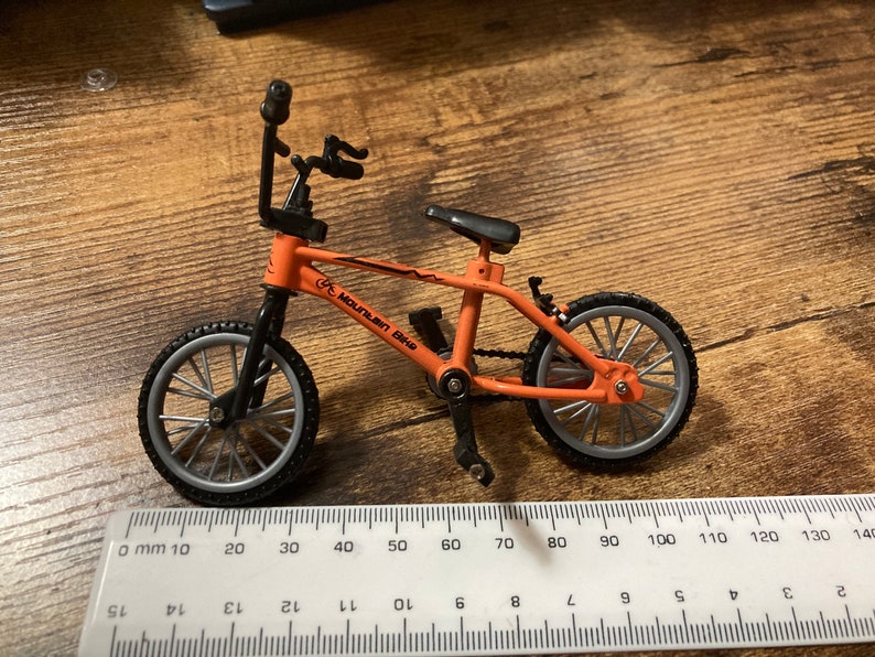 Op de afbeelding: Een oranje en zwarte miniatuur mountainbike met de woorden "Mountain Bike" op het frame. De fiets heeft zilveren wielen en een zwart zadel. Een liniaal staat op de voorgrond.