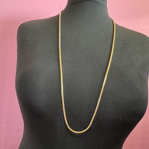 May include: A long, gold chain necklace on a black mannequin. The chain is a simple, snake-like design.
