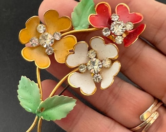 May include: A vintage floral brooch featuring three flowers in yellow, white, and red enamel, each with a sparkling rhinestone center. The gold-tone metal brooch includes green enamel leaves.