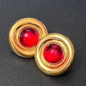 May include: A pair of gold tone earrings with red glass cabochon stones. The earrings are round and have a raised rim around the stones.