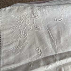 May include: White embroidered linen textile featuring a floral design. The embroidery showcases a detailed flower with delicate petals and leaves, along with trailing vines and decorative elements. The edges have a scalloped trim.