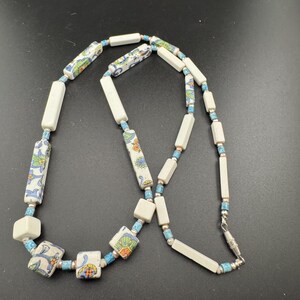 May include: A necklace with a mix of white, blue, and patterned ceramic beads. The necklace has rectangular and square beads, with floral designs on the patterned beads. The beads are strung together with a blue cord and silver-coloured metal clasps.