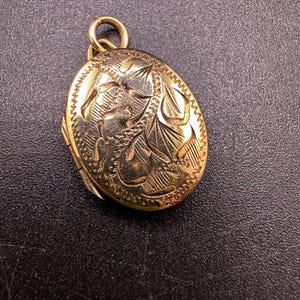 May include: An oval-shaped gold-toned metal locket featuring an intricate engraved pattern on its front surface. The locket includes a small circular jump ring at the top for attachment, with visible hinge details along the side of the casing.