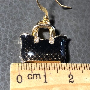 Puede incluir: Pendientes en forma de bolso de mano en tono dorado con esmalte negro y pedrer&iacute;a transparente. Los pendientes miden aproximadamente 1 cm de largo.
