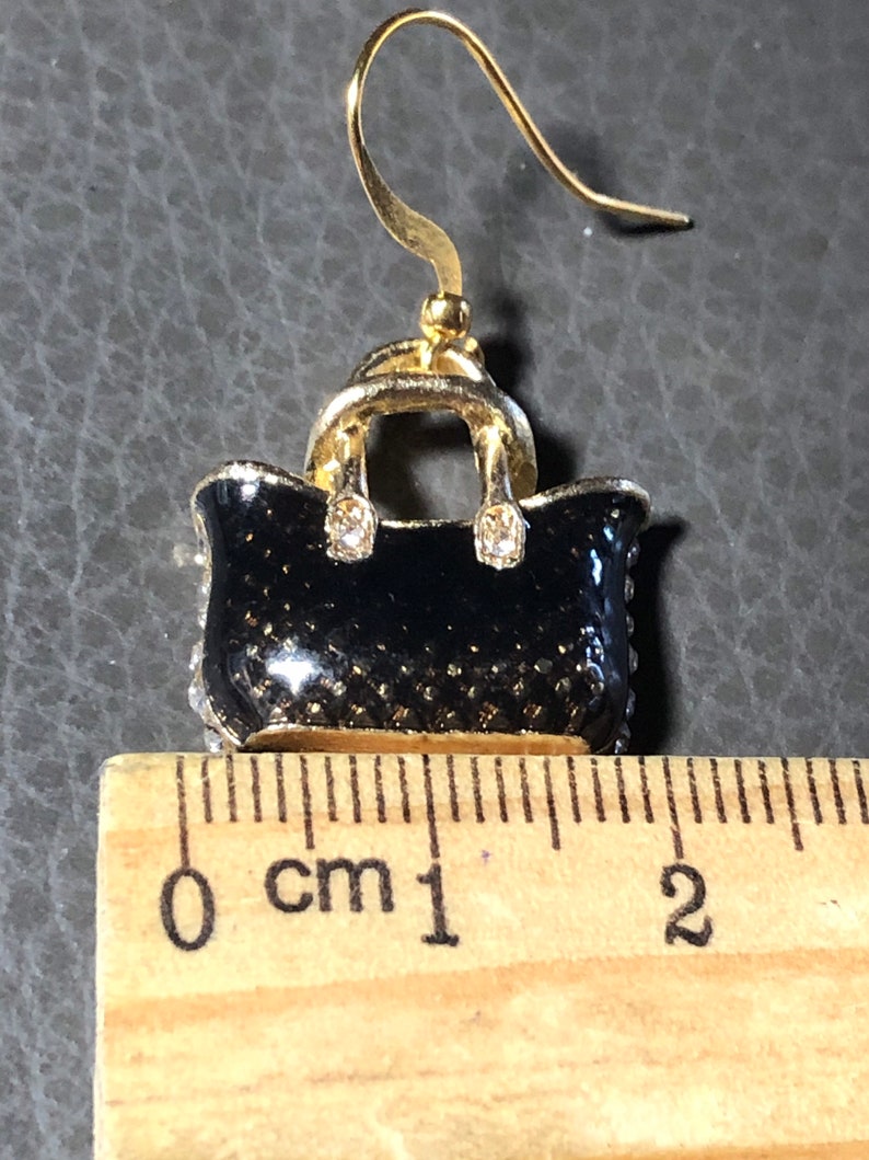 Puede incluir: Pendientes en forma de bolso de mano en tono dorado con esmalte negro y pedrer&iacute;a transparente. Los pendientes miden aproximadamente 1 cm de largo.