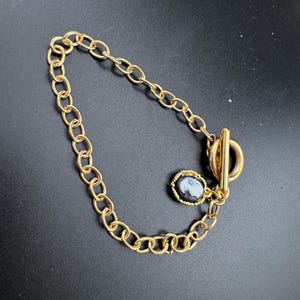 May include: Gold-coloured chain bracelet with a toggle clasp. The bracelet features a small, oval cameo pendant with a profile portrait in grey and black. The jewellery is displayed against a dark background.