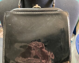 以下が含まれることがあります： 正方形で、上部に留め具が付いた、黒いエナメル革製のハンドバッグ。バッグには、上の部分にハンドルが2つ付いています。