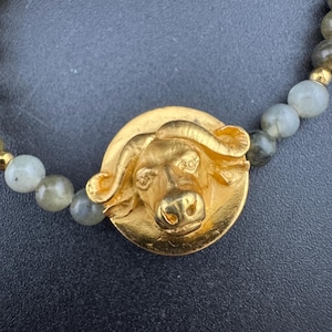 May include: A gold-colored bracelet featuring a detailed buffalo head pendant. The pendant is centered on a strand of gray and gold-colored beads, creating a striking contrast. The bracelet is set against a dark background.