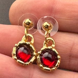 May include: A pair of gold earrings with red gemstone accents. The earrings have a simple chain design and a round, red gemstone in the center.