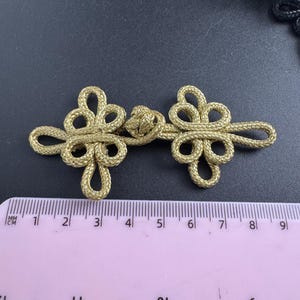 May include: Gold-colored Chinese knot button closure. The button is made of woven cord and features a symmetrical design with a central knot and two flower-like shapes. The button measures approximately 3 inches in length.