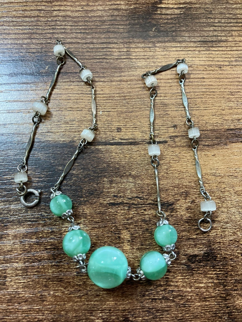 Pu&ograve; includere: Una collana a catena d'argento con una chiusura d'argento e perline di vetro verde. Le perline hanno un motivo a spirale e sono collegate da anelli d'argento.