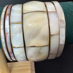 以下が含まれることがあります： 手作りのブレスレットのコレクション。ブレスレットは、白、クリーム、ピンクの色合いの長方形の真珠母貝のピースで構成されたモザイクデザインが特徴で、金色の金属フレームにセットされています。ブレスレットは緑色のベルベットの表面に展示されています。