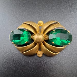 May include: Vintage gold-tone brooch with two large emerald-green oval-cut rhinestones. The brooch has a stylized floral design with curved, layered petals and small decorative accents. A classic piece of jewelry.