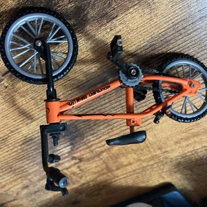 Op de afbeelding: Een oranje en zwarte miniatuur BMX-fiets met zilveren wielen. Op het frame van de fiets staat de tekst "MOUNTAIN BIKE". Dit gedetailleerde model is waarschijnlijk een speelgoed of verzamelobject.