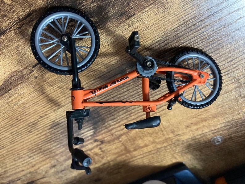 Op de afbeelding: Een oranje en zwarte miniatuur BMX-fiets met zilveren wielen. Op het frame van de fiets staat de tekst "MOUNTAIN BIKE". Dit gedetailleerde model is waarschijnlijk een speelgoed of verzamelobject.