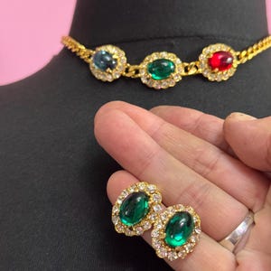 May include: A gold-coloured necklace and earring set. The necklace has three oval gemstones in blue, green, and red, each with small clear crystals. The earrings are oval green gemstones with a halo of clear crystals.