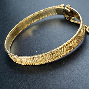 May include: A gold-colored bracelet with a textured, woven design. The bracelet features a decorative pattern along the center and a clasp closure. The jewelry is displayed on a dark surface.