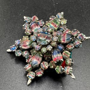 May include: A star-shaped brooch featuring a central round iridescent stone surrounded by marquise and round faceted crystals in shades of blue, green, and red. The stones are set in a silver-toned metal frame with a multi-layered, dimensional design.