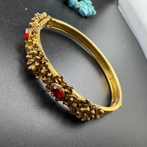 May include: A gold-tone hinged bangle bracelet featuring a textured, floral-inspired surface with raised metal details and two inset oval-shaped red gemstones. The bracelet has a smooth interior band and a secure clasp mechanism for closure.