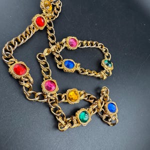 May include: Gold-tone chain necklace with colorful, round gemstones. The necklace features red, pink, yellow, green, and blue stones set in ornate, textured bezels. The chain has a classic curb link design.