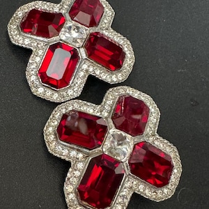 May include: A pair of red and clear rhinestone earrings in the shape of a cross. The earrings are silver-toned and have a geometric design.
